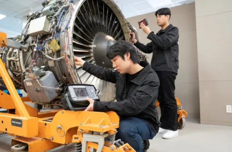 검은 실습복 차림 두 사람이 큰 엔진 앞에서 도구를 대고 있는 모습