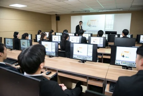 여러 사람이 컴퓨터 앞에 앉아 실습실 앞쪽 설명을 듣고 있는 모습