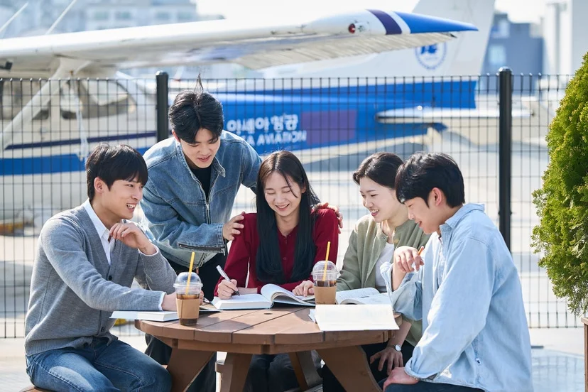 야외 둥근 탁자에 다섯 사람이 모여 자료와 음료를 놓고 대화하고 있는 모습
