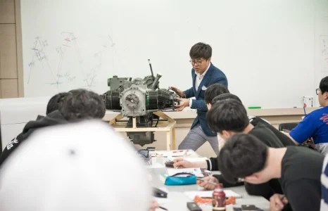 교사가 책상 위 엔진 모형을 두고 학생들에게 설명하는 모습