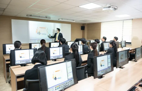 컴퓨터가 놓인 교실에서 여러 사람이 화면을 보고 앞에 선 사람이 설명하고 있는 모습