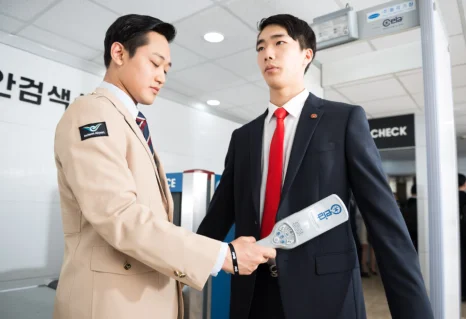베이지색 제복 차림 사람이 정장 차림 사람 앞에서 휴대용 검색기를 대고 있는 모습