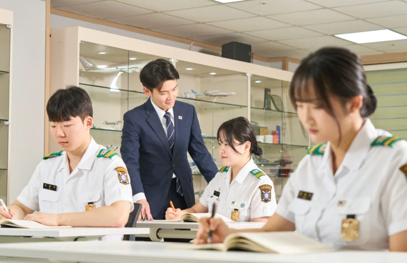 교실 책상에서 제복 차림 사람이 글을 쓰고 정장 차림 사람이 뒤에서 몸을 숙여 보고 있는 모습