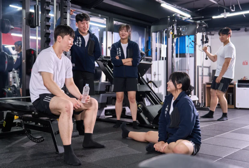 운동 시설에서 앉은 사람 주위로 여러 사람이 모여 설명을 듣고 있는 모습