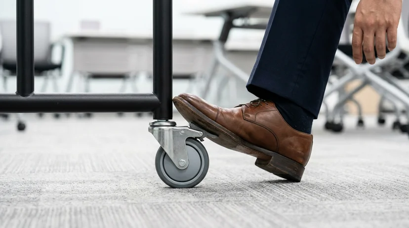 A technician's shoe pressing down on the locking mechanism of a wheel base at the bottom of a display stand.