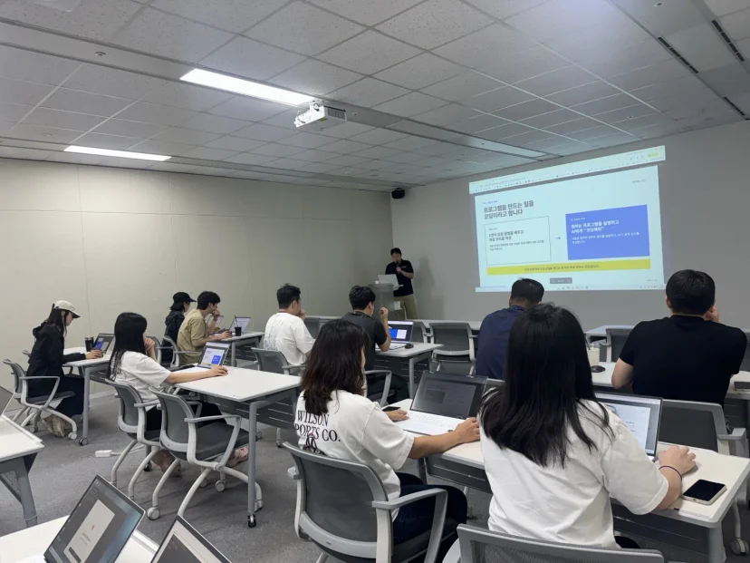 AI 응용 과정, 프로그램을 만드는 일을 코딩이라고 합니다 슬라이드 앞에서 강의하는 모습과 윌슨 티셔츠를 입은 수강생들