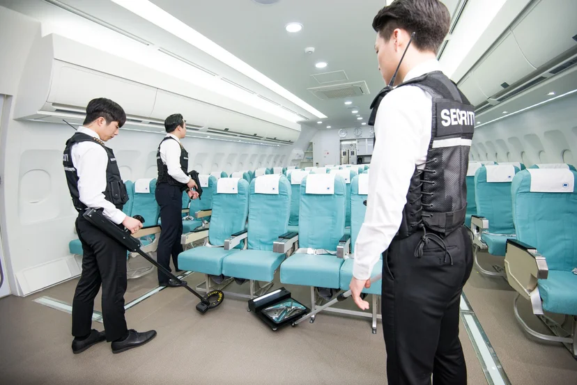 항공기 객실 모형에서 휴대용 탐지기로 좌석 주변을 검색하는 학생들