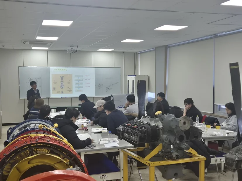 엔진과 부품이 놓인 실습 공간에서 함께 작업하는 사람들