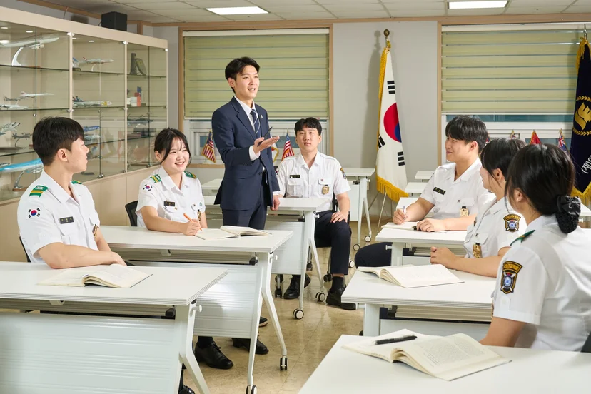 강의실 책상에 앉아 교수의 설명을 듣는 제복 차림 학생들