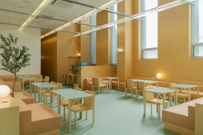 Shared lounge at Mangrove Dongdaemun with pale tables, wooden chairs and tall windows by a reception counter