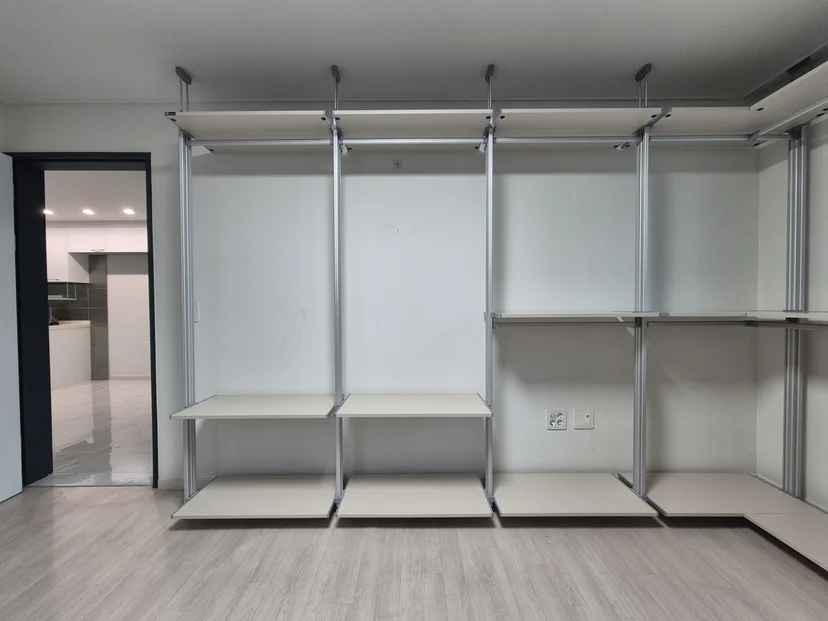 무지 순백 선반과 밝은 회색 옷봉이 천장에서 매달린 일자형 시스템행거. 실버 크롬 기둥이 수직으로 지지하며, 선반 2단과 상단 옷봉 영역이 명확히 보인다. 밝은 목재 바닥재와 흰 벽면 배경.