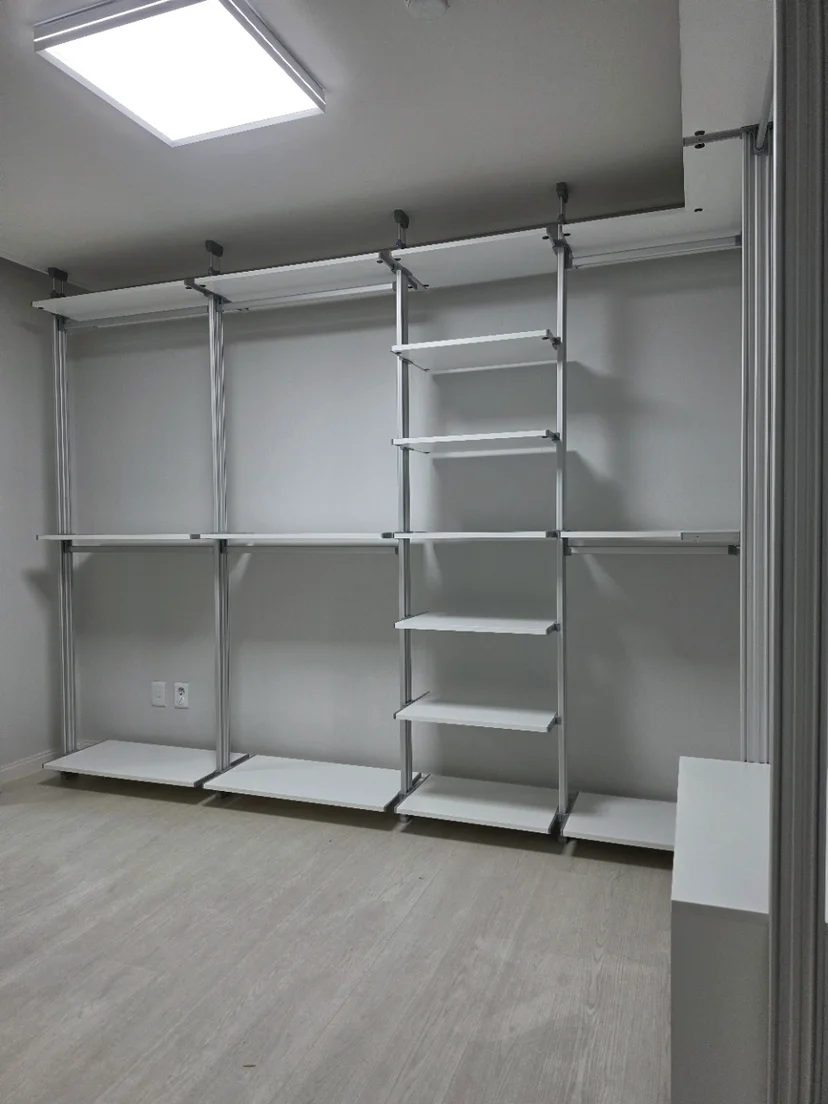순백 선반과 하부장으로 구성된 일자형 시스템행거. 실버 크롬 기둥이 명확히 보임. 옅은 우드톤 바닥재가 깔려있고 밝은 천장 조명.