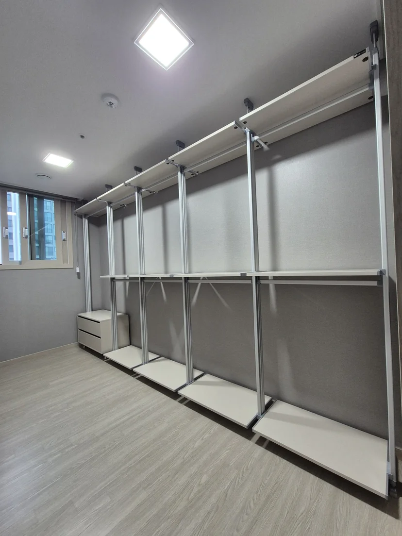 무지 순백 상판과 밝은 회색 하판 조합의 일자형 행거 3개 유닛. 상단에 옷봉, 중단과 하단에 선반 배치. 실버 크롬 기둥. 좌측에 흰색 4단서랍장. 연목조 바닥.