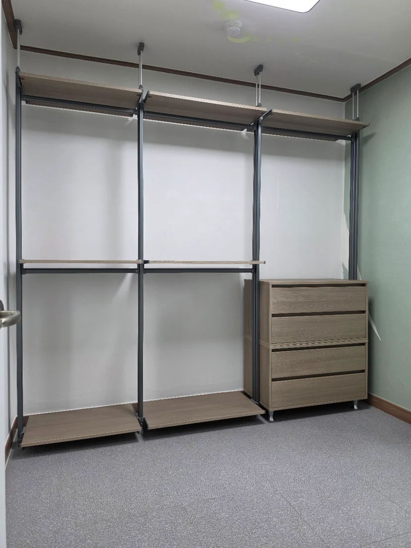 밝은 목재 선반과 짙은 적갈색 4단서랍장이 배치된 드레스룸. 검정 기둥과 크롬 옷봉이 상단에 설치되어 있고, 회색 카펫 바닥에 나무 베이스가 놓여 있다.