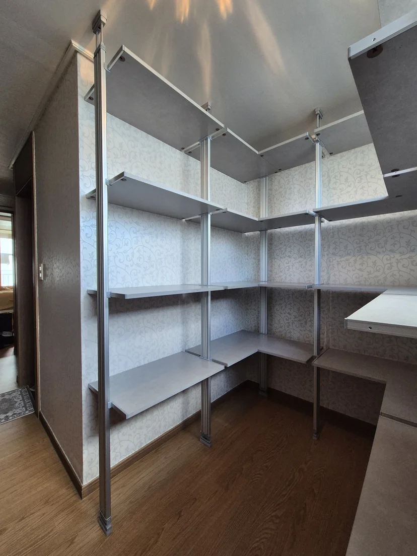 밝은 회색 콘크리트 선반과 크롬 실버 기둥으로 구성된 ㄱ자형 시스템행거. 두 벽면에 걸쳐 4단 선반과 상단 옷봉이 설치돼 있으며, 바닥은 갈색 목재.