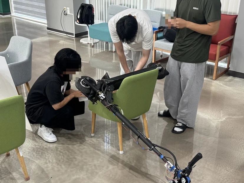 킥보드를 의자에 걸쳐 놓고 함께 배선을 점검하는 팀원들