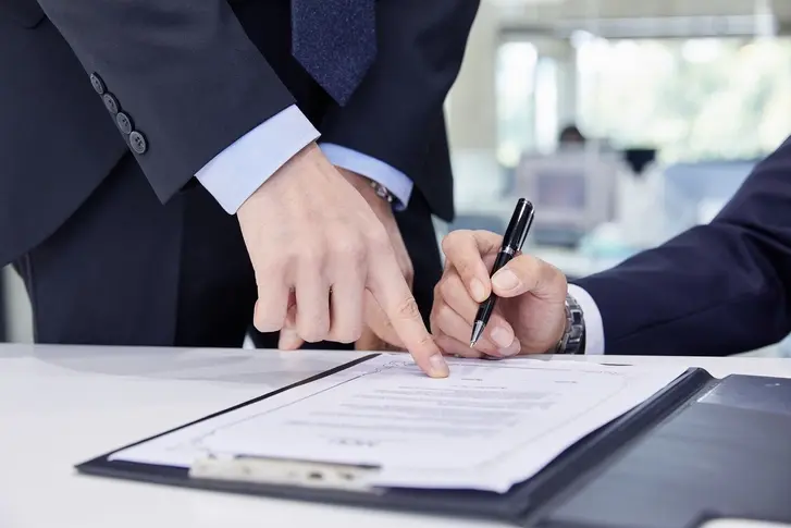 검지손가락으로 서류의 서명란을 가르키는 사람과 서명하기 위해 펜을 잡고 있는 사람의 손이 보이는 이미지