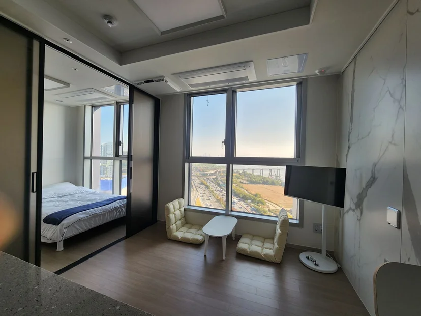 Studio living area with lounge chairs, a TV and a Han River view through large windows, adjoining glass-walled bedroom (Listing ID : 32655)